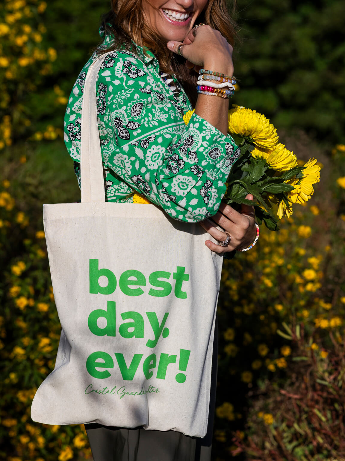 A woman outdoors holding a cream canvas tote bag with green text reading 'best day. ever!' and 'Cecel Greenhouse' below it, while holding yellow flowers.