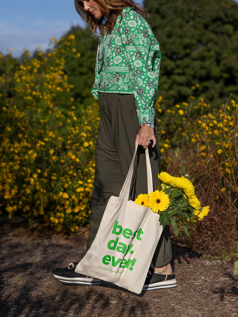A person standing outdoors in a field of yellow flowers, wearing a green patterned long-sleeve top and dark olive pants, holding a cream canvas tote bag with green text that reads 'best day ever!' and a bouquet of yellow flowers.