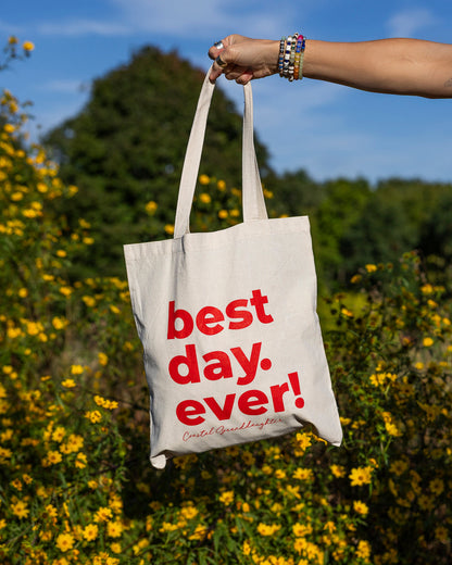 A cream canvas tote bag with bold red text reading 'best day. ever!' is held up by a person's arm in a sunny outdoor setting filled with yellow flowers and green trees.