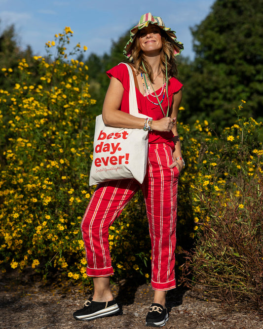 A woman stands outdoors in a field of yellow flowers wearing a red and white striped shirt and matching pants, a green hat, and carrying a white tote bag with red text that reads 'best day. ever!'.