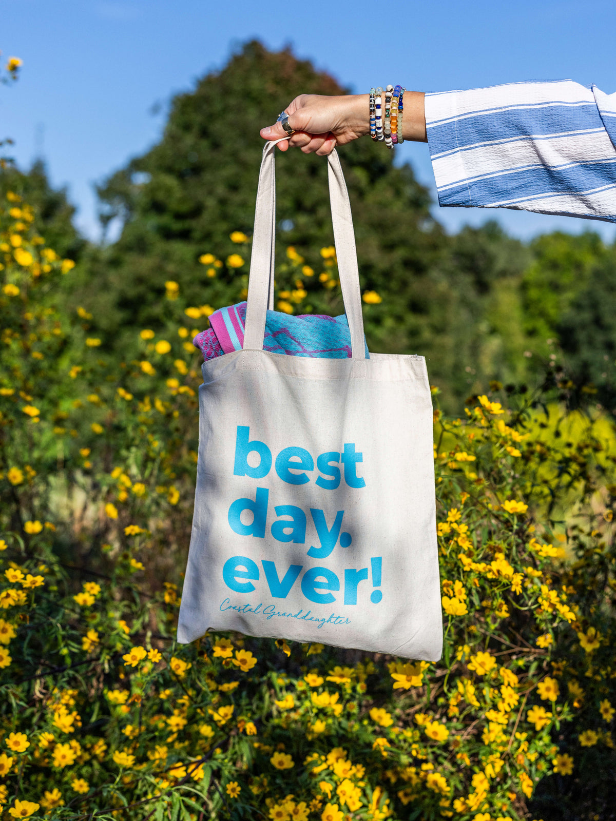 A person's hand holding a cream canvas tote bag with blue text that reads 'best day. ever!' against a background of yellow flowers and green trees.