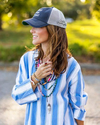A woman outdoors wearing a blue and gray trucker hat with white embroidered text, a blue and white vertically striped button-up shirt, and multiple layered beaded necklaces.
