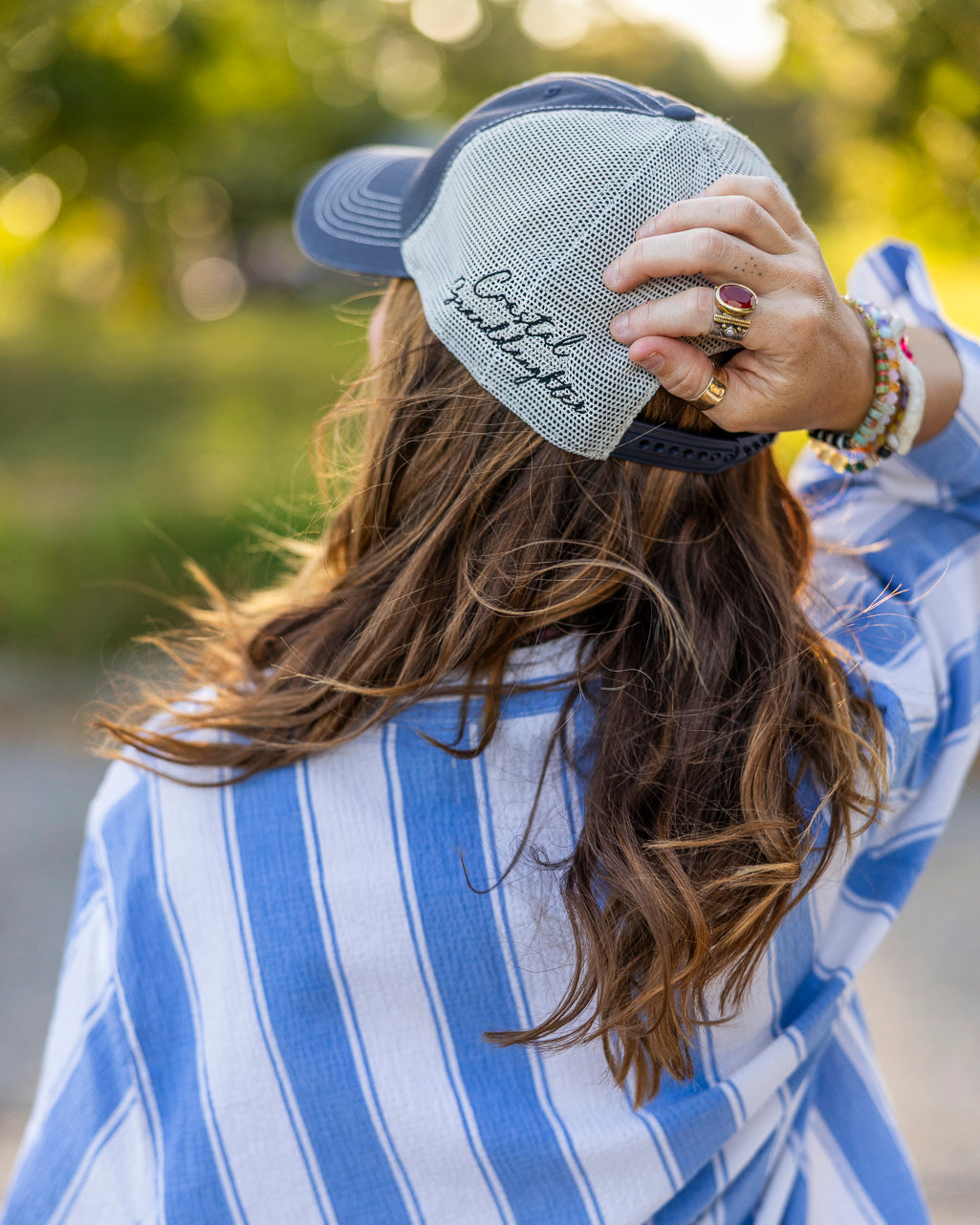 A woman seen from behind wearing a blue and white striped shirt and a gray mesh baseball cap with 'Coastal Granddaughter' embroidered on the side, set against a blurred green outdoor background.