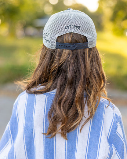 A person viewed from behind wearing a light gray mesh-back baseball cap with 'EST 1980' embroidered on the back, along with a blue and white vertically striped shirt, set against a blurred outdoor background.